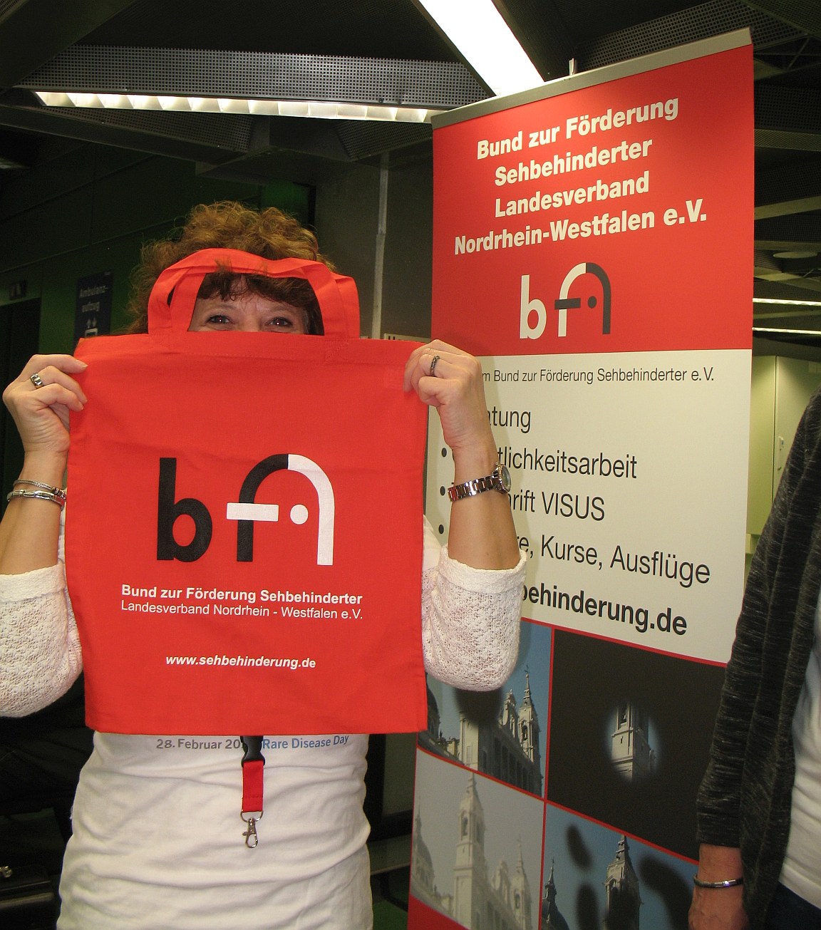 Elisabeht Krych an einem BFs-NRW-Infostand mit Vereins-Rollup hält sich eine BFS-NRW-Stofftasche vors Gesicht