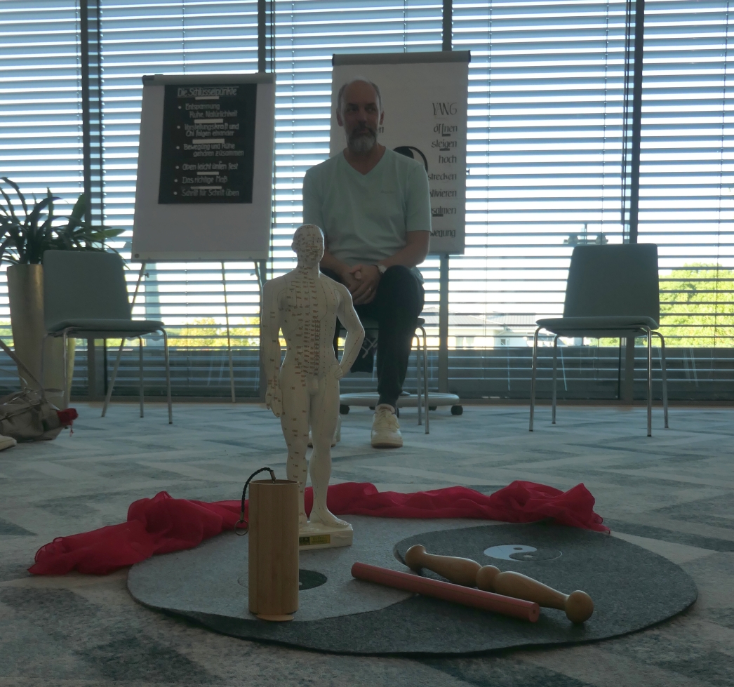 Der Kursleiter seitz auf einem Stuhl und erklärt, davor auf einem Yin-Yang-Teppich eine Akupressur-Puppe und Holzstäber